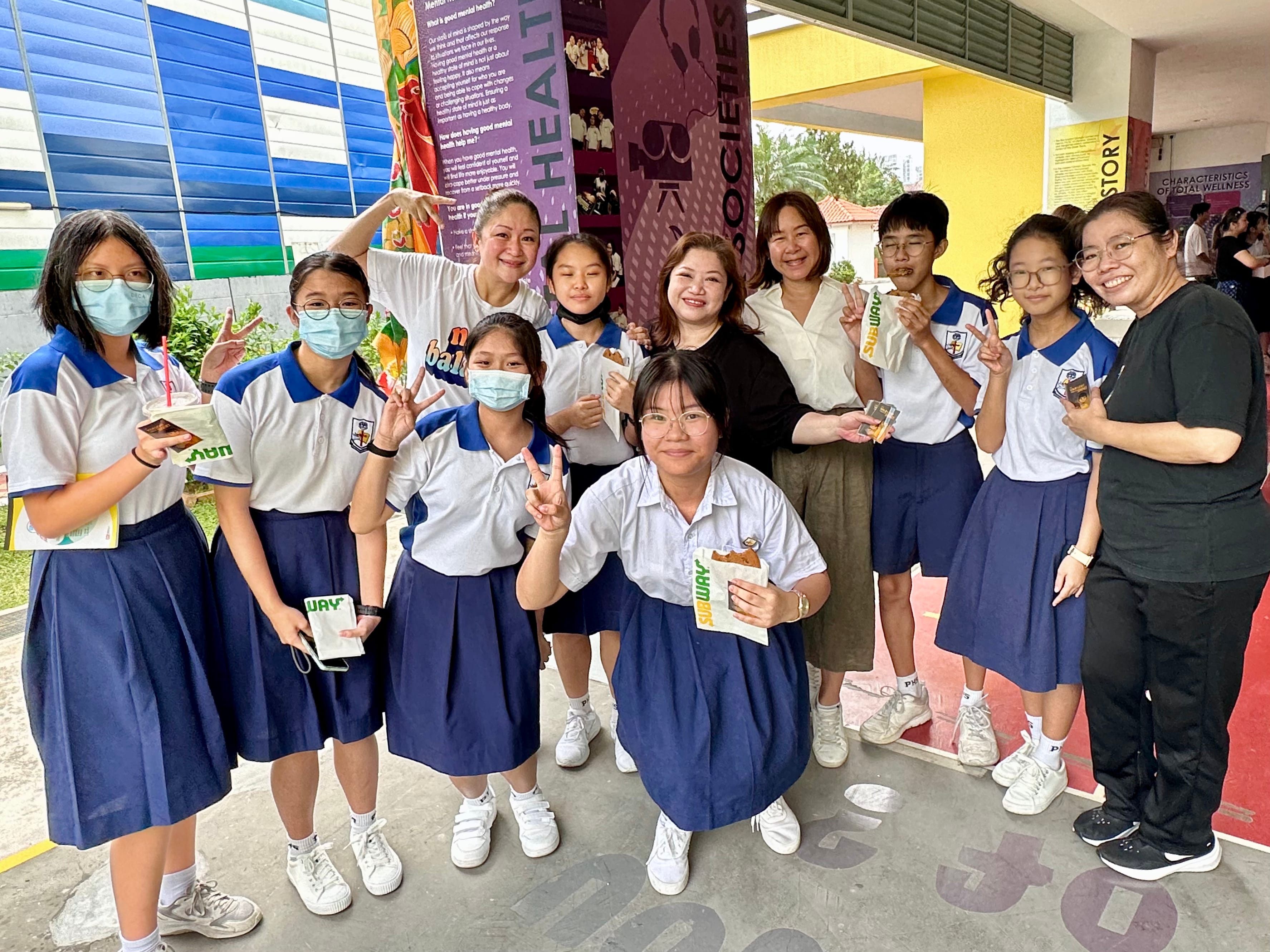 Group of students in school uniforms and teachers posing with peace signs, some holding Subway snacks.
